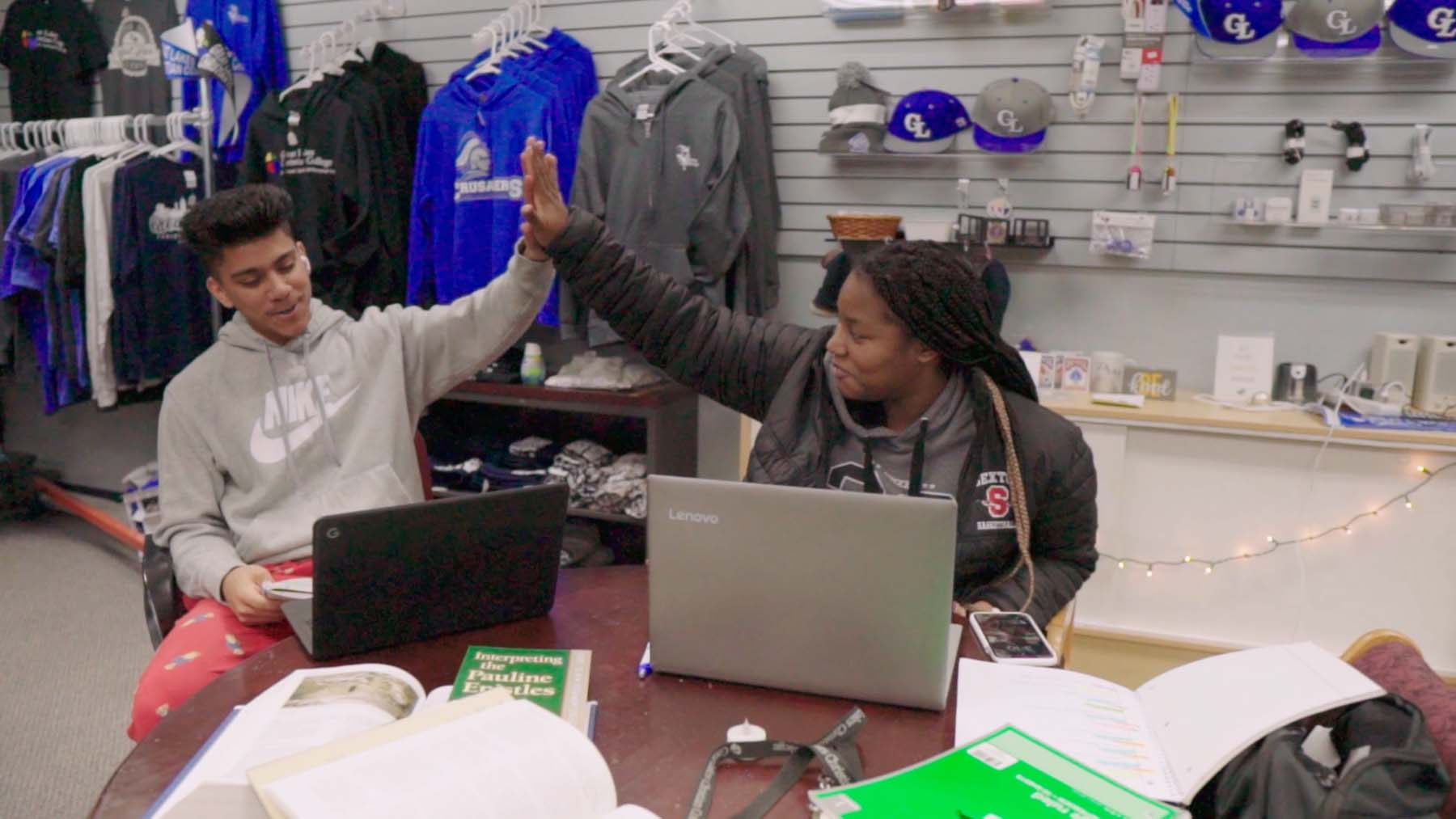 students sitting at a table in the bookstore studying on laptops giving each other a high five